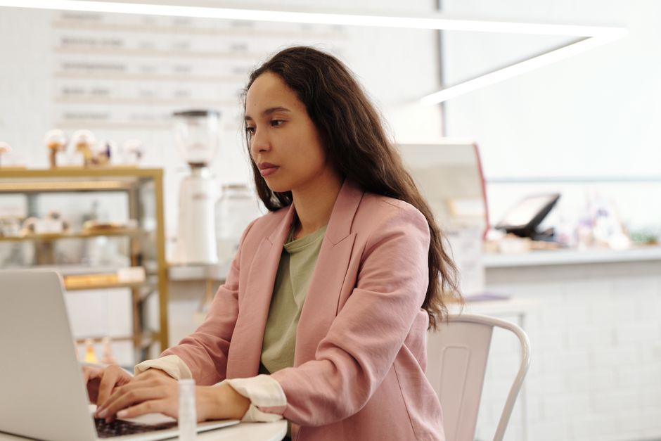 A young woman typing on a laptop beside coffee in a bright cafe
